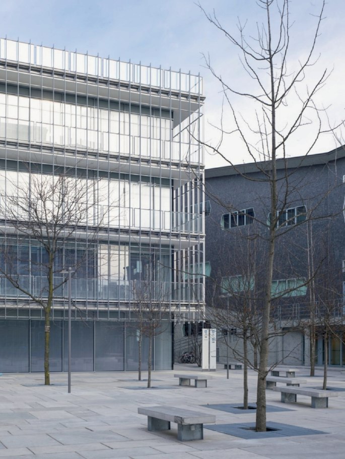 A photograph of the exteriors of a glass building. Multiple benches are randomly arranged near the trees.