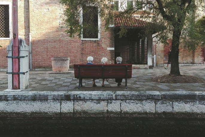 A photograph of a street shows a bench with three elderly people sitting on it. There is a large tree and a house nearby.