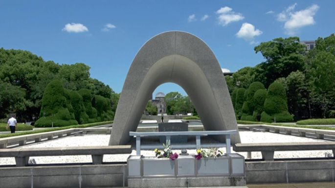 A photograph of the Hiroshima Peace Memorial. It has an arch construction in the center that is surrounded by a parapet wall. Flower bouquets are arranged in the center. The area is surrounded by dense forests.