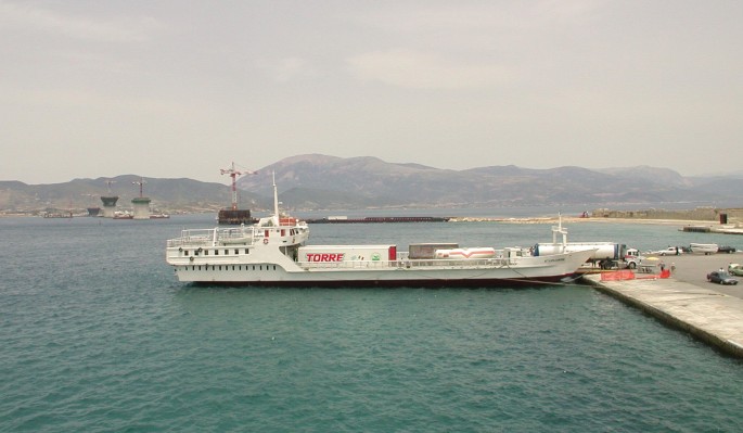 A photograph of the Rio-Antirio bridge which is under construction over the water body. There is a passenger ship on the water body and a hill station in the background.