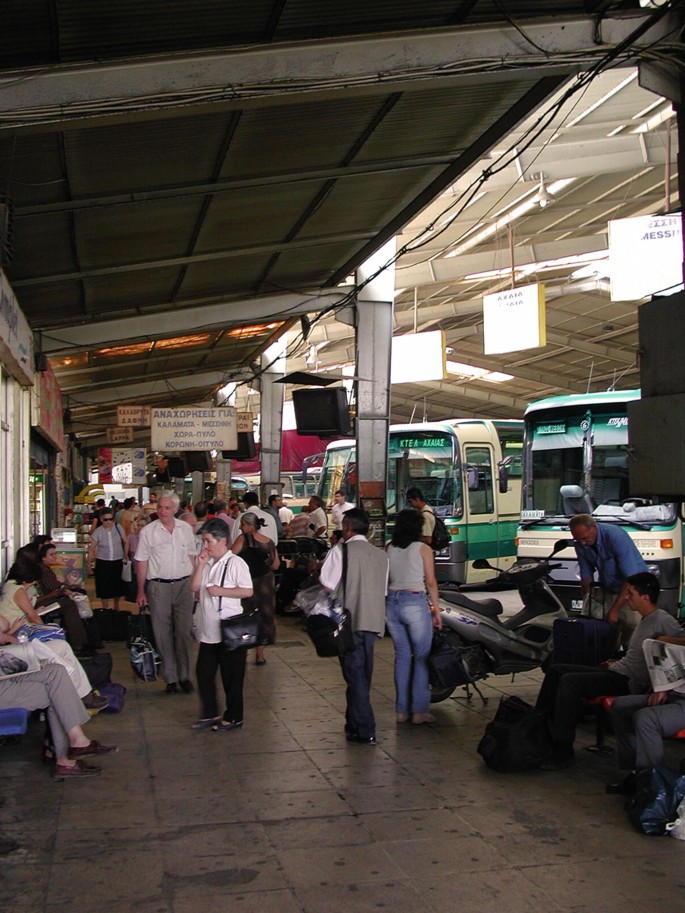 A photograph captures the bustling scene at Kifissou Station, depicting people strolling, seated on chairs with their luggage, and the presence of various vehicles, including two-wheelers and buses parked in the vicinity.