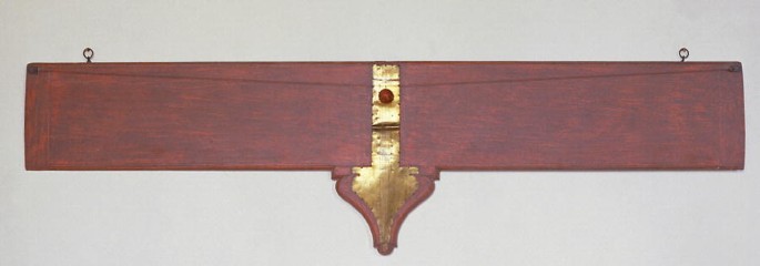 A photo. A rectangular wooden block with 2 pegs on either end is suspended tightly from 2 nails on a wall. A central, narrow, and shiny vertical strip extends down to an inverted onion-shaped base.