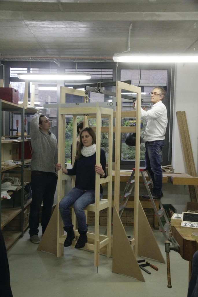 A photo. A woman sits on a Sanctorian chair's prototype while 2 men, 1 on the left, and another on a small conical ladder on the right, examine it standing. The chair has 2 rectangular supporting structures on either side and is suspended from a horizontal bar with a weight on the opposite end.