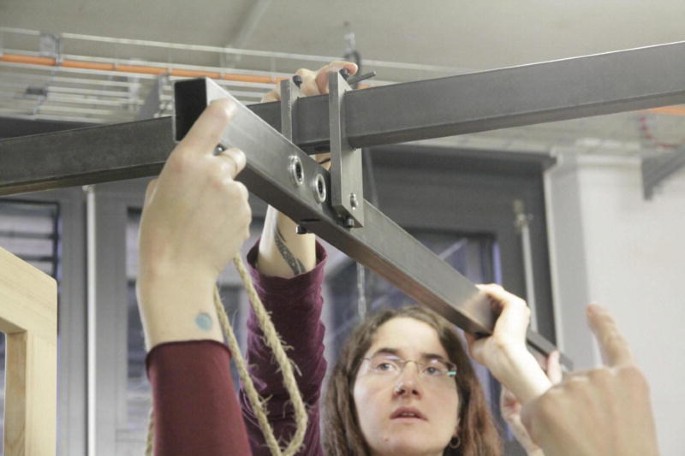 A close-up of the suspension of the horizontal bar from the pivot. A woman holds one side of a bar above her head at the back while another hand supports it from the foreground. It reveals 2 ball bearings arranged in a sequence.