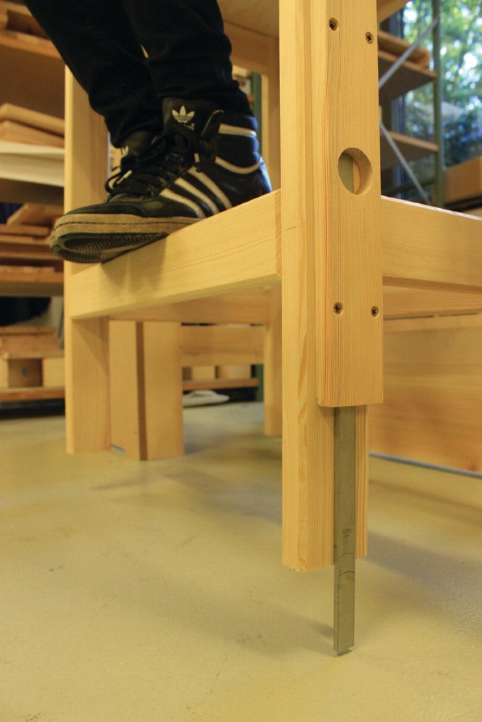 A photo. An oblique view from below of the Sanctorial chair's prototype. A pair of legs wearing a shoe appears on the top of the chair. A vertical steel bar is attached to the leg closest to the camera, and it extends down to the floor, with the actual leg lifted above the floor.