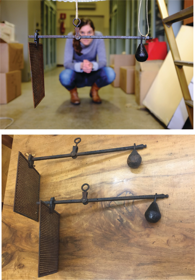 2 photos. 1. A replica of the water measurement setup hangs from above, in the foreground. A woman gazes at it from the background. 2. An overhead view of a pair of replicas placed on a floor. Both have iron plates, bars with pegs, and weights, from left to right, in order.