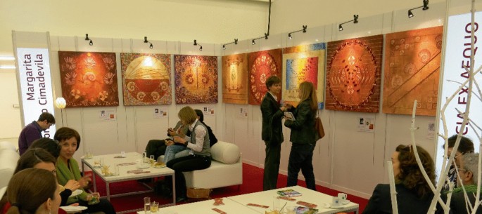 A photograph of a group of people sitting in an arts and science exhibition with posters and paintings of murals on the walls. It reads texts in a foreign language.