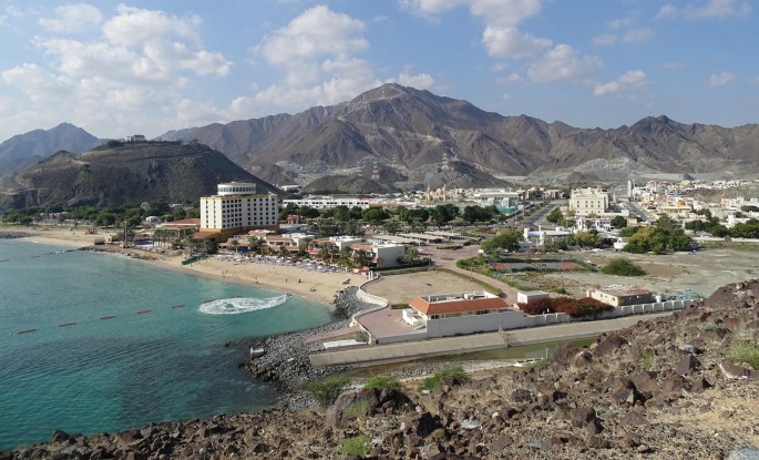 A photo depicts the coastal development in Khor Fakkan. There are huge buildings and roads, along with mountains in the background.