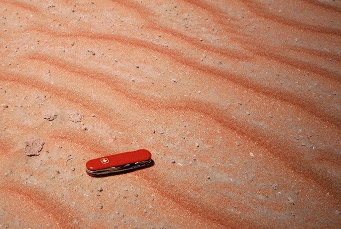 A photograph of a Swiss knife placed on a sandy, wavy surface