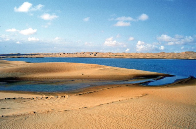 A photograph of a sandy desert with a body of water in the background. The water is calm and reflects the blue sky above. There are some small waves lapping against the shore, and the sand is dotted with small puddles of water.