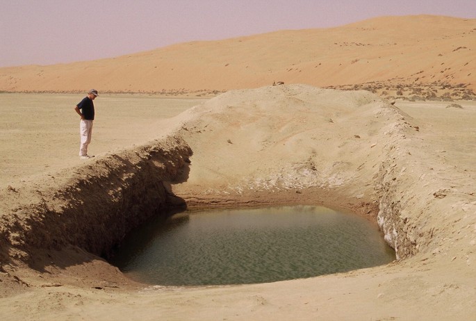 A photograph of a man standing on the top of the edge of a large water hole in the ground. The hole is surrounded by sand and dirt, and there is a small pool of water at the bottom of the hole. The man is wearing a black shirt and black pants, and he is looking down into the hole.