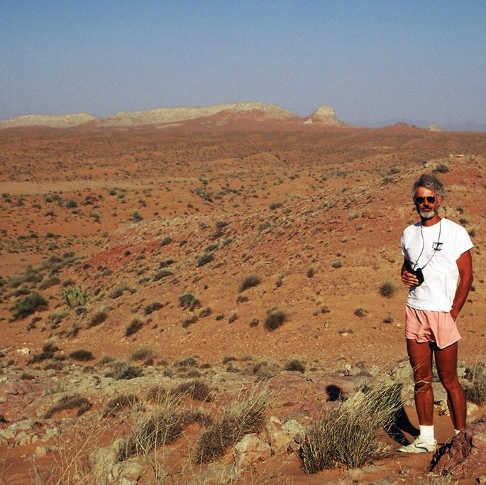 A photograph of a man standing on a desert. He is wearing a white t-shirt and shorts. In the background, there are mountains and a blue sky. There are some shrubs and rocks in the background.