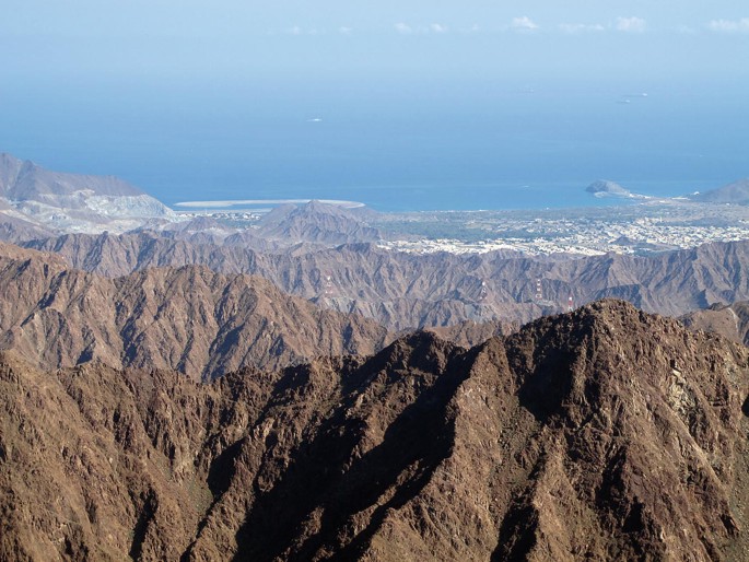 A photograph of a view of a mountain range with a body of water in the background. The mountain range is made up of brown and tan rock formations, with some areas covered in green vegetation. The body of water is a deep blue color and appears to be a large bay or inlet.