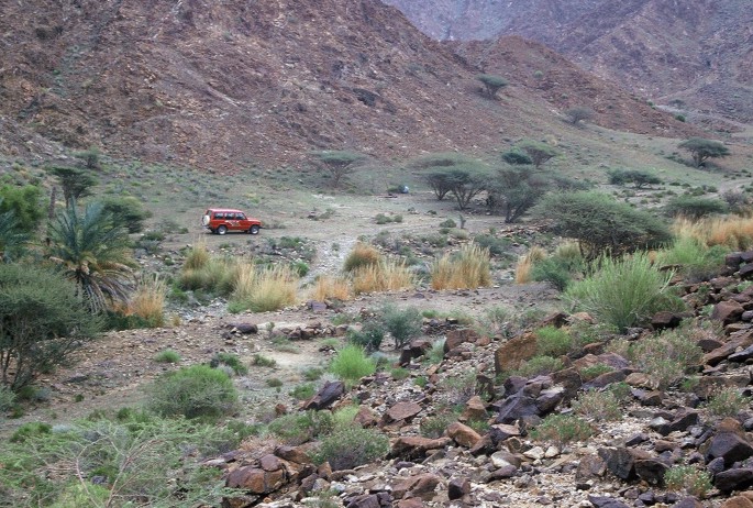 A photograph of a rugged mountain landscape with rocky terrain and vegetation. There is a small dirt road winding through the landscape, and a red car parked on the side of the road.