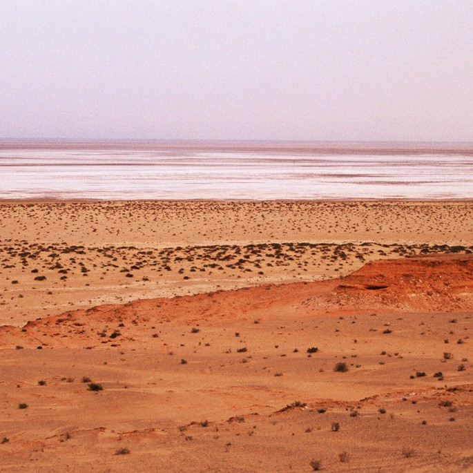 A photograph of a large body of water in the background with sand dunes in the foreground. The sky is clear and there are no clouds in sight. The photograph appears to be taken from a high altitude, giving a bird’s eye view of the landscape.