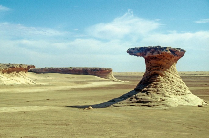 A photograph of a large rock formation in the middle of a desert. The rock formation is made up of different shapes and sizes, with some parts being jagged and others being smooth. The rock formation is surrounded by sand and dirt, with some small plants growing on the surface. The sky is clear and blue, with a few white clouds in the distance.