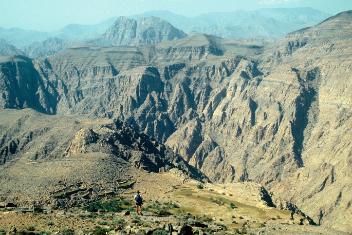 A panoramic view of a man standing before towering, rugged mountains.