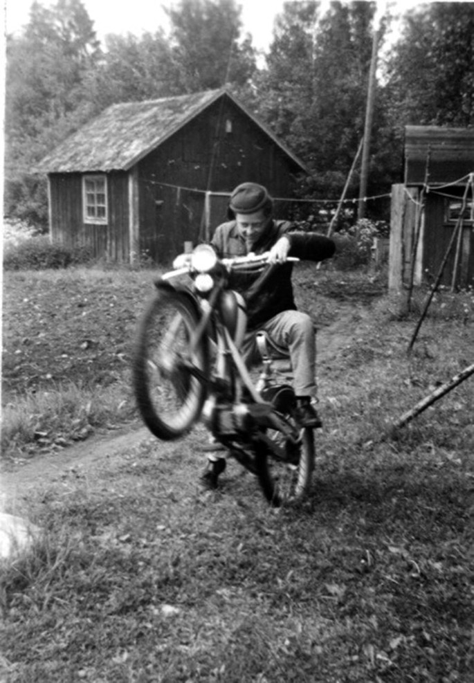 A photo of a man on a motorbike in a landscape of thick vegetation. The front wheel is raised high at an angle as he supports himself with his right leg on the ground. The left leg is on the footrest, and he looks down. A small hut is in the background.