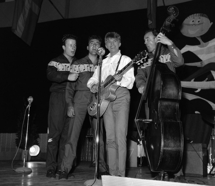 A still from a band performing on stage. 4 men stand on stage and sing into a mic. The one at the center plays a guitar and the one to his left plays a cello.