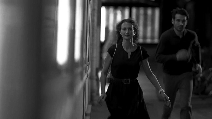 A grayscale photo of a scene in a video. It has a woman running forward, followed by a man running behind.