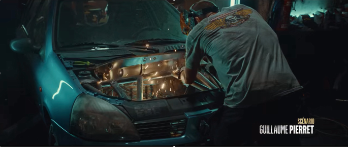 A screenshot of a movie scene presents the backside of a man welding in the car's engine space.