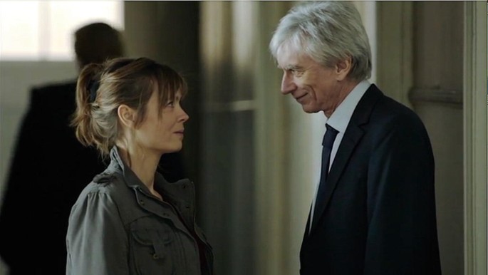 A screenshot from a video series, titled Engrenages. It displays the female character, Laure, and a male character, Roban, exchanging a smile facing each other.