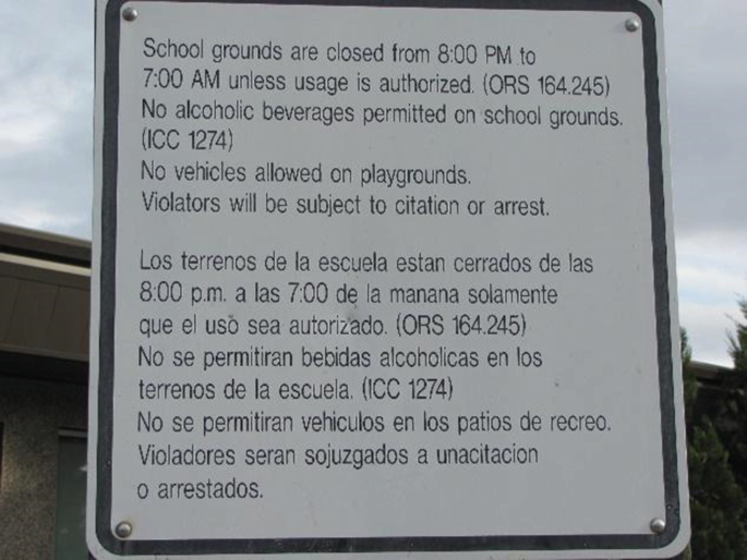 A photo of a sign has text that reads, school grounds are closed from 8 P M to 7 A M unless usage is authorized. No alcoholic beverages permitted on school grounds. No vehicles allowed on playgrounds. Violators will be subject to citation or arrest. Translation in a foreign text appears below.