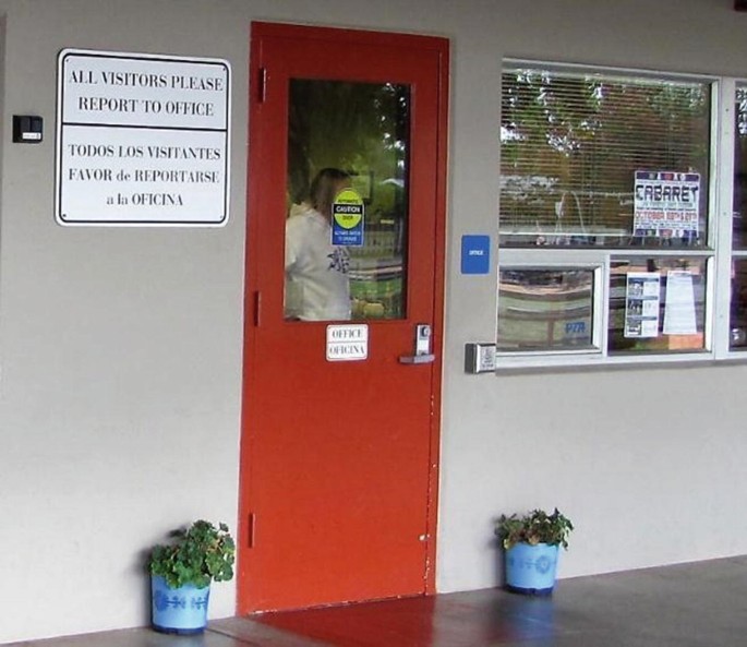 A photograph of the entrance of a building that has several signs on it. A large sign on the left of the door reads all visitors please report to office. Translation in a foreign language appears below it. 2 small signs on the door read, caution, and office. A poster on the window reads, cabaret.