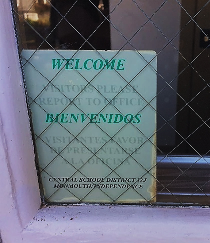 A photo of signage behind a metal grill window reads, welcome, visitors please report to office. The translation in a foreign language appears below it. Beneath it more text reads, central school district 13 J, Monmouth independence.