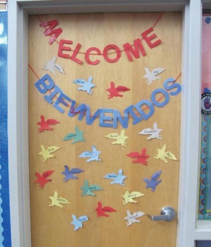 A photo of a display on a door that has letters strung together to spell the word welcome. Another string of letters spells out a translation of the same in a foreign language. The door is decorated with shapes of birds in flight with some text written on them.