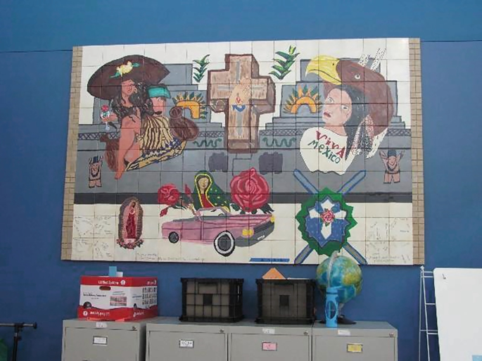 A photo of a large canvas made of smaller square pieces stuck together, which has been displayed on a large wall. The canvas features a painting of Mexican motifs, including 2 women wearing ornate hats, a 3 D cross, roses, a large fort, and a shield with swords. Text on 1 woman reads, viva Mexico.
