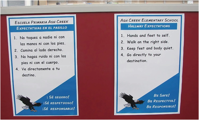 A photo of 2 posters displayed on a wall. The poster on the right reads, Ash Creek elementary school, hallway expectations. Instructions include hands and feet to self, walk on the right side, and go directly to your destination. A translation in a foreign text is on a poster on the left.