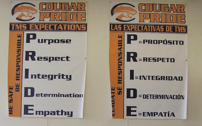 A photo of 2 posters displayed on a wall. The poster on the left is titled Cougar pride, T M S expectations. An acrostic for the word Pride is featured. On the left of the acrostic, a slogan reads, be safe, be responsible. A poster on the right has a translation of the same in a foreign language.