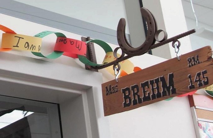 A close-up photo of a sign made from a wooden plank with jagged edges. It hangs from 2 hooks attached to an ornate metal frame that is fixed above a door. Text on the sign reads, Mrs. Brehm, R M 145. A paper chain with handwritten text on it, has been draped over the sign.