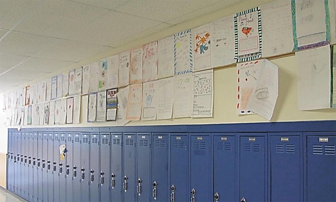 A photograph of the walls of a school hallway. The wall above the lockers is filled with large hand drawn posters, which have been pasted side-by-side in 2 long rows that stretch the entire length of the hallway. The posters contain drawings and text. A few posters hang in a crooked position.