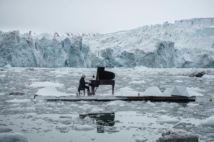 A photograph of a person sitting on a small platform in the middle of a body of water surrounded by icebergs. The person is wearing a black suit and is playing a piano.