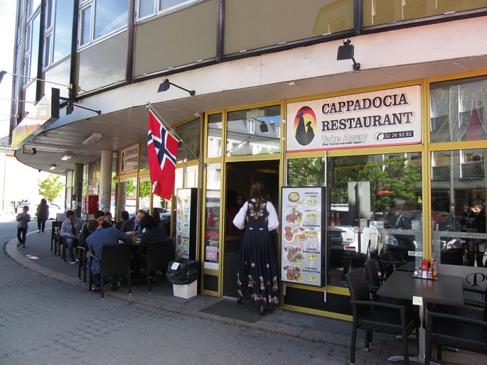 A photograph captures the inviting entrance of a restaurant, bustling with activity as numerous patrons enjoy their meals at outdoor tables and chairs.