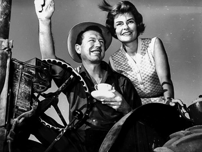 A photo of Nico Carstens and Vera Gibson. He sits on a vintage cabriolet behind a steering wheel. He holds a cup and saucer on his left hand and waving his right hand and smiles wide gazing to the right. Vera Gibson stands next to him and smiles.