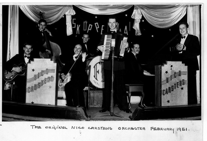 A photo of a 7-member orchestra troop performing on stage. It includes men playing guitar, violin, cello, organ, and accordion in the front. A man plays drums at the back, seated behind these men. 2 tall desks on either side bear the text, Nico Carstens.