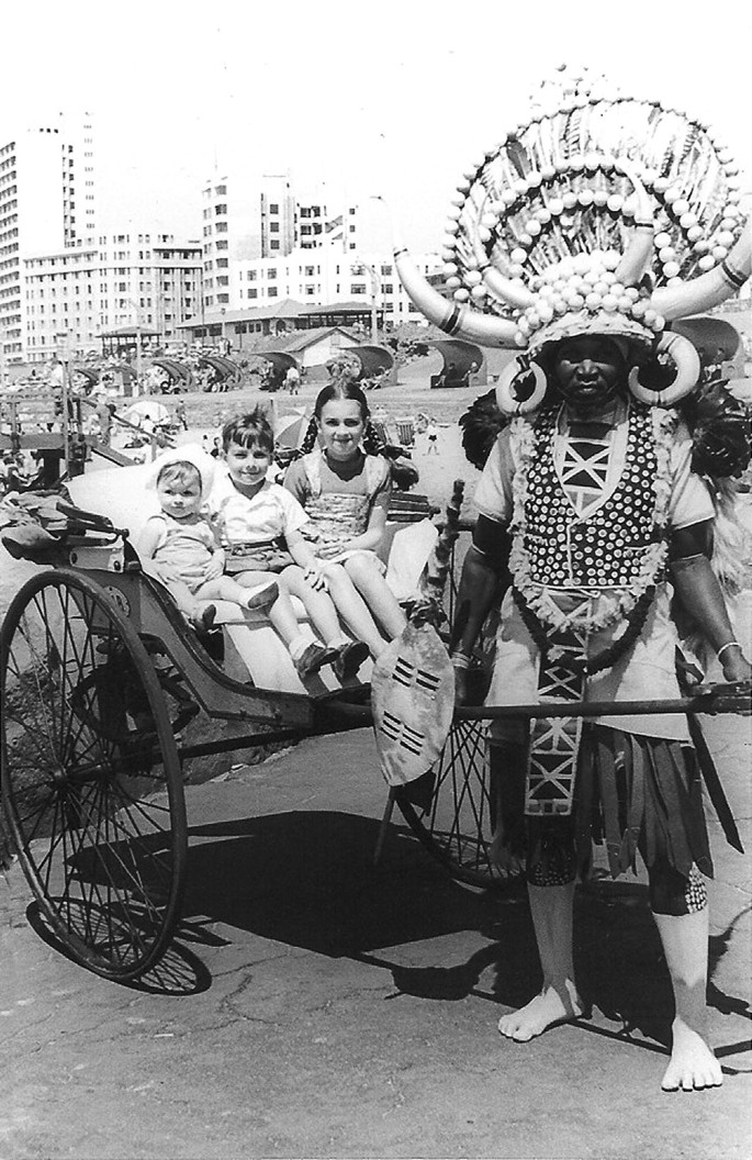 A photo of a rickshaw-puller. He stands posing for the camera holding the front lever of a rickshaw with his hands. 3 children of different ages are seated on the rickshaw. The rickshaw puller wears an elaborate head gear with animal horns and feathers and is dressed in traditional tribal attire.