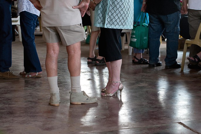 A photo of a pair of several human legs in different attires and gender assembled in an indoor space. A pair of legs sporting a pair of short trousers and shoes and another sporting pointed heels and knee-length shorts face each other in the foreground.