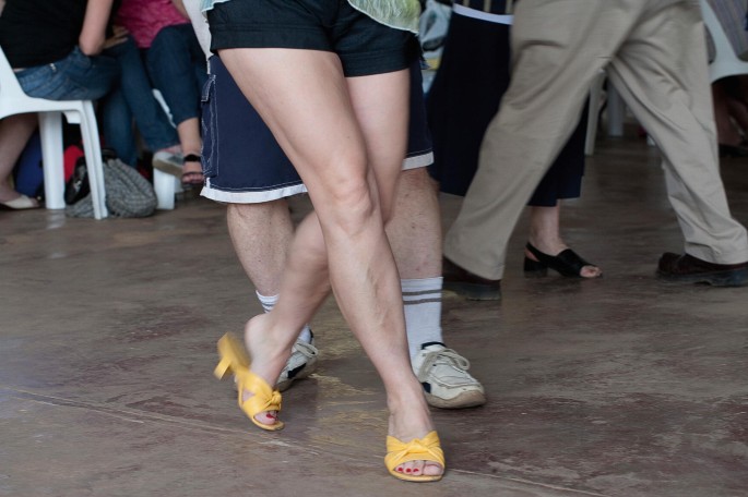 A photo of a pair of 2 legs in close-up view. A pair sporting slip-on sandals, polished nails, and short trousers poses a crisscrossed stance. The pair behind in sneakers and knee-length trousers follows the direction of the formers stance.