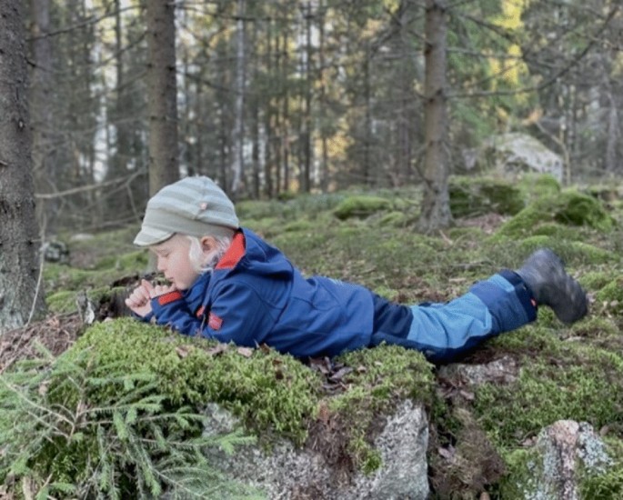 A photo of a child lying down on the ground. The background depicts a thick growth of trees.