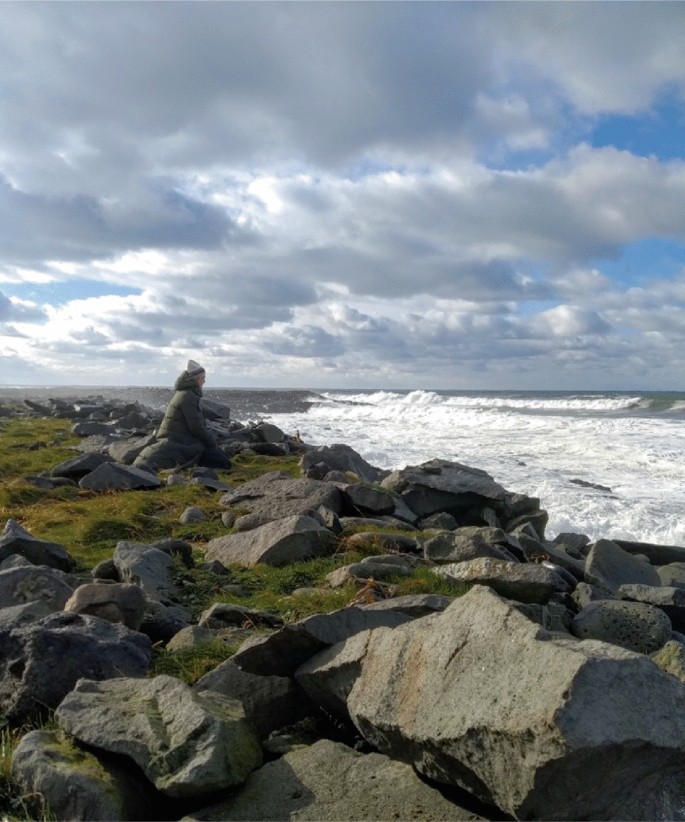 A photo of a person sitting on the rocky shore of the sea watching the frothy waves.