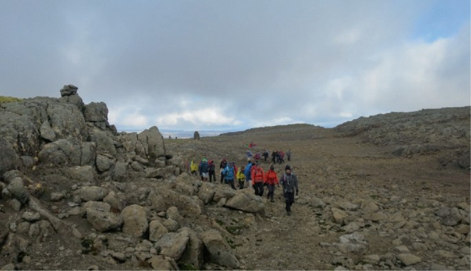 A photograph of a group of people trekking on a rocky mountain. The people walk near a mound of rocks.