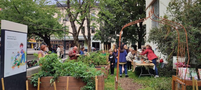 A photo of a small open cafeteria space within an urban apartment complex. It has a vertical row of plants cultivated in troughs. Some women are engaged in serving food at a small table on the right while some men and women eat from a small plate held in their hands.