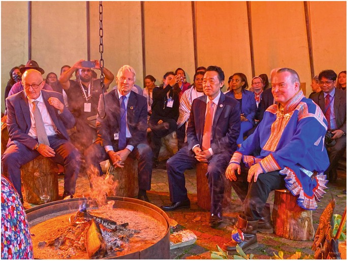 A photo of 4 people around a fire sitting on tree stumps. They are from right to left Anders Oskal, Qu Dongyu, Morten von Hanno Aasland, and Miguel Garcia Winder. There are people seated behind them.