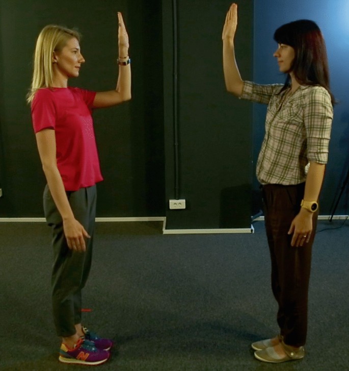 A photograph of women engaged in a mirror task, while facing each other. On the left, a woman stands and raises her left hand, while on the right, another woman stands and raises her right hand.