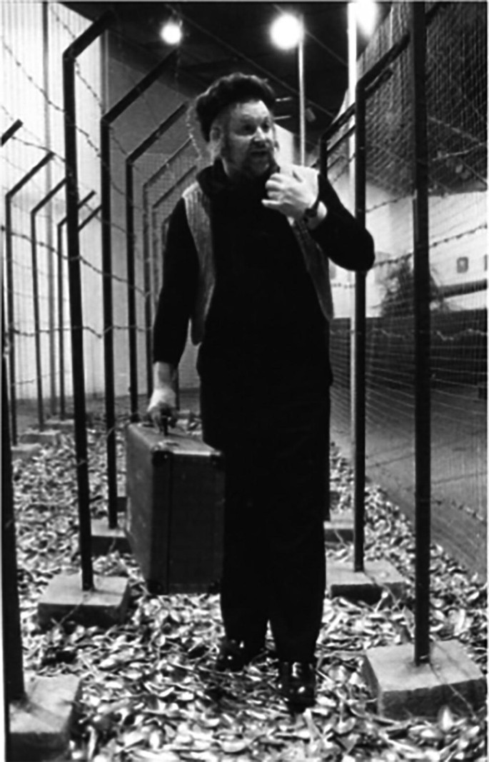 A photograph of Wolf Vostell walking through a fenced area with a suitcase in his hand.