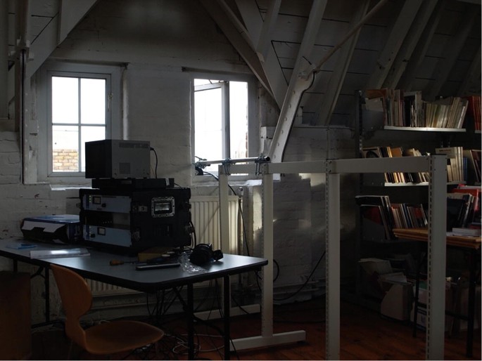 A photograph of the room with the equipment on the table, a chair near the table, and a bookshelf in the right corner.